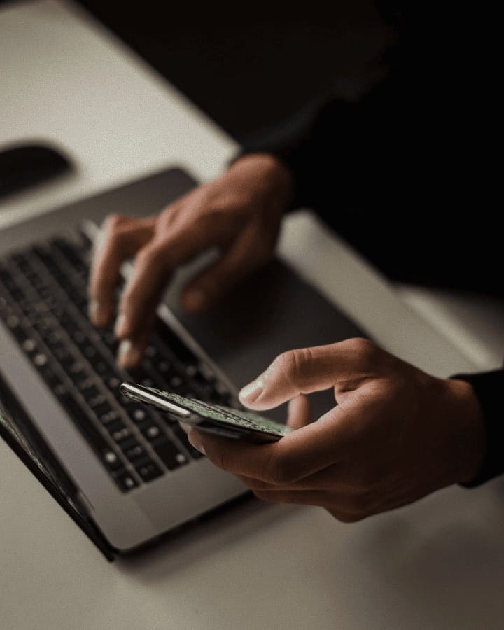 Someone with one hand on a macbook, while the other is holding a phone, seemingly reading something.